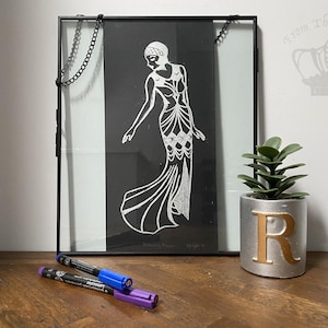 May include: A black framed print with a white Art Deco illustration of a woman in a gown. The image is on a wooden surface, with a small plant and two markers. The frame has a chain.