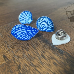 May include: Four blue and white seashell-shaped enamel pins. The pins feature detailed designs of various seashells. One pin is attached to a white backing with a silver pin back. The pins are displayed on a wooden surface.