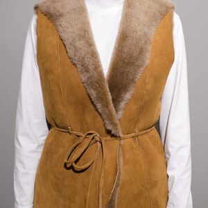 May include: A brown suede vest with a faux fur collar and a tie belt. The vest has a long, fitted style and is perfect for layering.