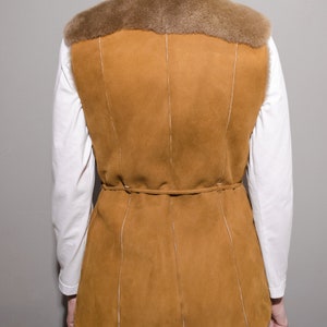 May include: A brown suede vest with a fur collar and a tie belt. The vest has a long, fitted style and is lined with a soft, brown fabric.