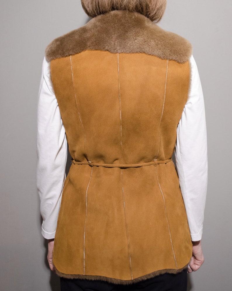 May include: A brown suede vest with a fur collar and a tie belt. The vest has a long, fitted style and is lined with a soft, brown fabric.