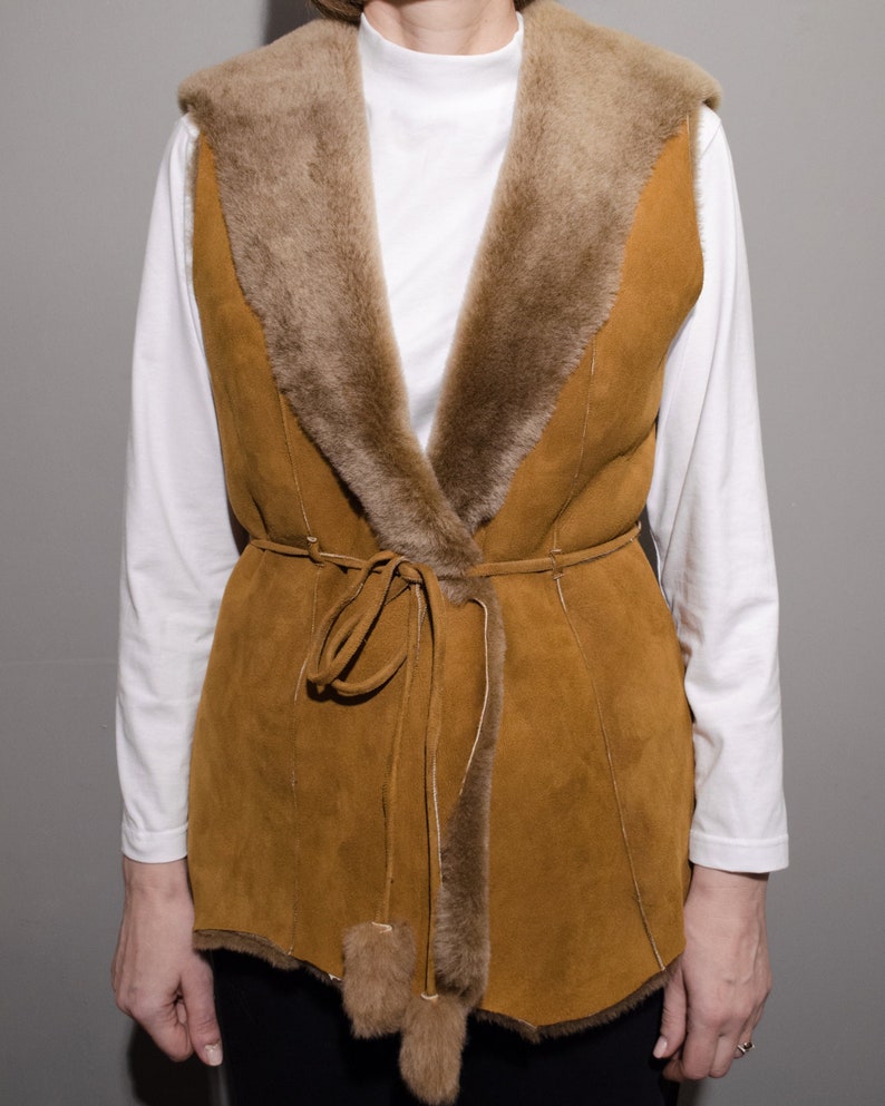 May include: A brown suede vest with a faux fur collar and tie closure. The vest has a long, fitted style and is perfect for layering.