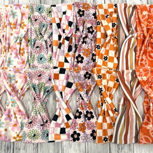 May include: A collection of colorful headbands with various patterns, including flowers, checkerboard, stripes, and peace signs. The headbands are in shades of pink, orange, black, and white, and are made of soft fabric.