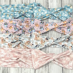 May include: Four colorful headbands with a knotted design. The headbands feature a repeating pattern of cartoon dogs and flowers on a light blue, pink, and white background. The bottom headband has a pink and white checkered pattern with paw prints.
