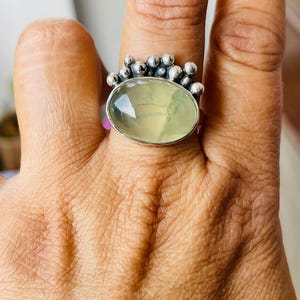 May include: A silver ring featuring an oval, pale green gemstone. The ring has a cluster of small, round silver accents around the top of the gemstone. The ring is worn on a finger, showcasing the design and color.