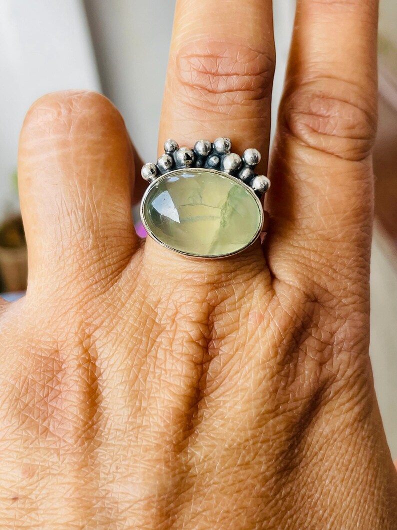 May include: A silver ring featuring an oval, pale green gemstone. The ring has a cluster of small, round silver accents around the top of the gemstone. The ring is worn on a finger, showcasing the design and color.