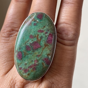Puede incluir: Un anillo de forma ovalada que presenta una gran piedra cabujón pulida con una base de color verde moteado, intercalada con distintas inclusiones de color rojo rosáceo intenso y vetas marrones finas, engastado en una banda de metal plateado.
