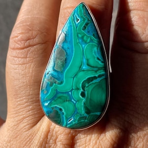 May include: A teardrop-shaped ring featuring a polished malachite and chrysocolla gemstone with vibrant swirls of deep forest green and bright turquoise blue, set within a smooth, simple silver metal band that frames the stone's natural patterns.