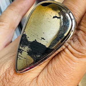 May include: A teardrop-shaped ring featuring a polished metallic pyrite stone with natural black inclusions, set in a smooth silver-toned metal band that wraps around the finger, showcasing a reflective surface and distinct mineral texture.