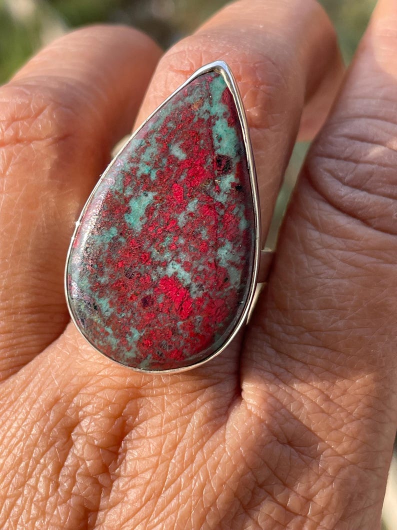 May include: A silver ring with a large, teardrop-shaped stone. The stone has a striking pattern of red and teal colours, set in a polished silver setting. This statement ring showcases a unique gemstone, perfect for adding a touch of colour.