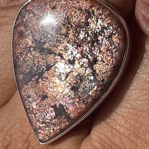 Puede incluir: Un anillo de piedra de sol en forma de lágrima con una gema pulida multicolor que presenta inclusiones metálicas brillantes en tonos cobre, oro y rosa, engastada en una banda de metal plateado lisa y sencilla que enmarca la superficie irregular.