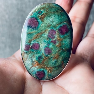 Könnte beinhalten: Ovale Ring mit einem großen, polierten Edelstein. Der Stein hat eine gesprenkelte grüne Basis mit roten Einschlüssen und eine silberne Lünette. Der Ring ist ein Statement-Stück, das natürliche Steinmuster zeigt.