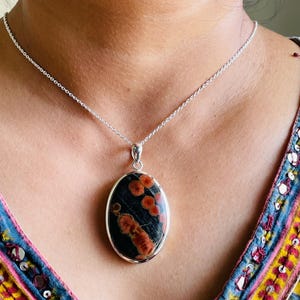May include: Oval pendant necklace featuring a dark stone with red and orange patterns, set in a silver-colored metal frame. The pendant hangs from a delicate silver chain, creating a striking contrast.