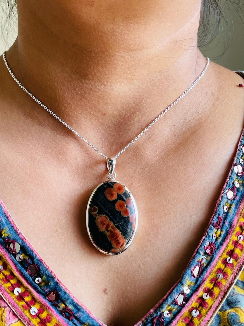 May include: Oval pendant necklace featuring a dark stone with red and orange patterns, set in a silver-colored metal frame. The pendant hangs from a delicate silver chain, creating a striking contrast.