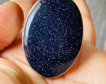 May include: An oval ring with a dark blue, speckled stone set in a silver-coloured metal band. The stone has a polished surface with reflective qualities. This piece of jewellery features a striking, dark blue gemstone.