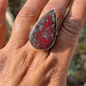 May include: A silver ring with a large, teardrop-shaped stone. The stone displays a mottled pattern of red and green colours, set within a silver band. The ring is shown on a hand.