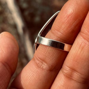 May include: A silver ring featuring a large, dark gemstone. The ring's band is smooth and polished, and the gemstone is oval-shaped. The ring is displayed on a finger, with a blurred background.