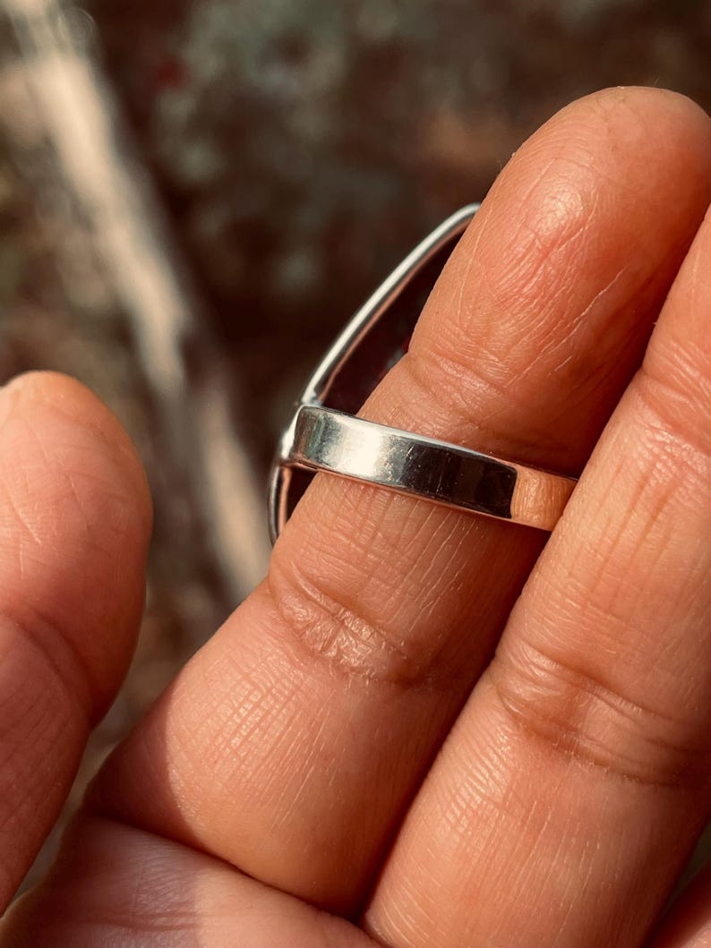May include: A silver ring featuring a large, dark gemstone. The ring's band is smooth and polished, and the gemstone is oval-shaped. The ring is displayed on a finger, with a blurred background.