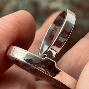 May include: A silver ring featuring a rectangular, reddish-brown stone. The ring's band is polished silver, and the stone has a mottled appearance. The ring is held in a hand, highlighting its design. The ring is a piece of jewellery.