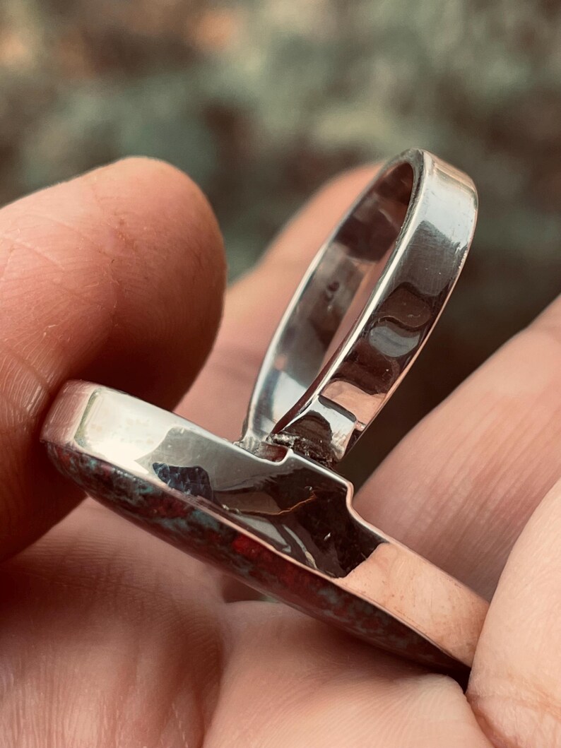 May include: A silver ring featuring a rectangular, reddish-brown stone. The ring's band is polished silver, and the stone has a mottled appearance. The ring is held in a hand, highlighting its design. The ring is a piece of jewellery.