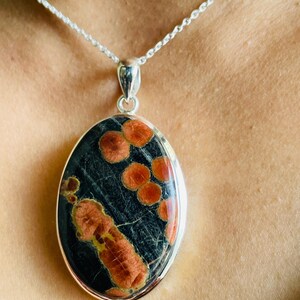 May include: Oval pendant necklace with a silver chain. The pendant features a dark background with orange and yellow patterns. The pendant is framed in silver.