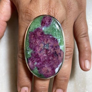 Könnte beinhalten: Ovaler Ring mit einem großen, polierten Edelstein. Der Stein zeigt eine lebendige Farbmischung mit einem zentralen Bereich in tiefem Magenta und umliegenden Grün- und Weißtönen. Die Fassung des Rings ist aus poliertem Silber.