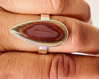 Könnte beinhalten: Ein tropfenförmiger Ring mit einem polierten Stein, der ein wirbelndes Muster aus Braun-, Burgunder- und Cremefarben aufweist. Der Stein ist in einem silbernen Band gefasst. Der Ring ist ein Schmuckstück.