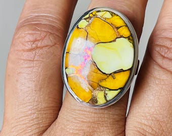 May include: A silver ring with an oval-cut stone. The stone displays a striking pattern of yellow and gold hues, with areas of lighter yellow and iridescent flashes. The ring has a plain, polished silver band, showcasing the stone's colours.