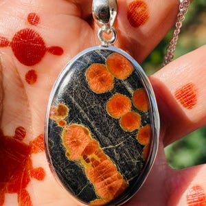 May include: Oval pendant necklace with a silver chain. The pendant features a black stone with orange and yellow patterns. The stone is set in a silver bezel. The necklace is displayed on a hand with red henna designs.