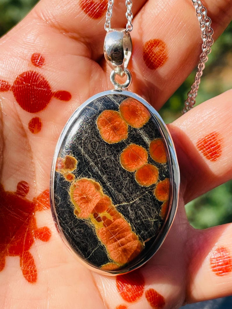May include: Oval pendant necklace with a silver chain. The pendant features a black stone with orange and yellow patterns. The stone is set in a silver bezel. The necklace is displayed on a hand with red henna designs.