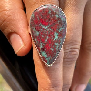 May include: A statement ring with a large, teardrop-shaped stone. The stone displays a mix of red and teal hues, set in a silver band. The ring is worn on a finger, highlighting its bold design and colour combination. The ring is a piece of jewellery.