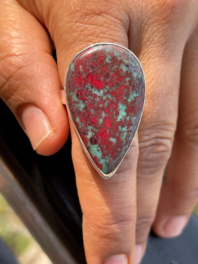 May include: A statement ring with a large, teardrop-shaped stone. The stone displays a mix of red and teal hues, set in a silver band. The ring is worn on a finger, highlighting its bold design and colour combination. The ring is a piece of jewellery.