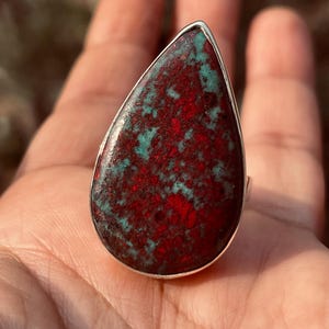 May include: A teardrop-shaped ring with a large stone displaying a striking red and teal pattern. The stone is set in a silver band. This statement ring is ideal for adding a pop of colour to any outfit.