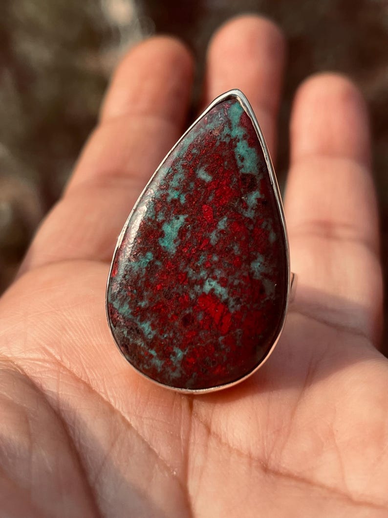 May include: A teardrop-shaped ring with a large stone displaying a striking red and teal pattern. The stone is set in a silver band. This statement ring is ideal for adding a pop of colour to any outfit.