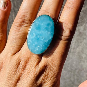 May include: An oval-shaped, light blue gemstone ring set in a polished silver-toned metal band. The stone features a smooth, opaque surface with subtle white cloud-like inclusions throughout, creating a marbled appearance across the entire face.