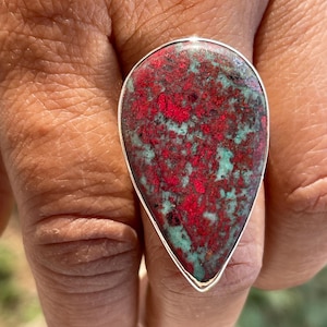 May include: A silver ring with a large, teardrop-shaped stone. The stone displays a striking pattern of red and teal hues. This ring is a piece of jewellery, suitable for fashion or personal adornment.