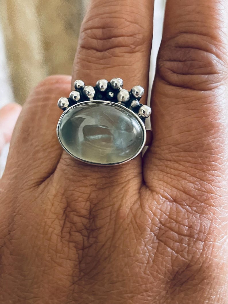 May include: Silver ring featuring a large, oval, pale green gemstone. The ring has a decorative silver band with small, round embellishments clustered around the gemstone. The ring is worn on a finger.