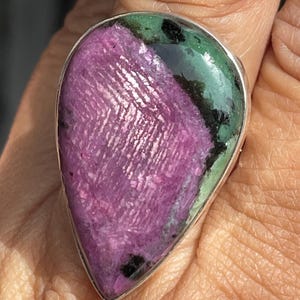 Puede incluir: Un anillo en forma de lágrima con una piedra preciosa de rubí zoisita pulida engastada en una banda de metal plateado. La piedra muestra un centro rosa magenta vibrante con inclusiones minerales verdes y negras, con una textura cristalina natural.