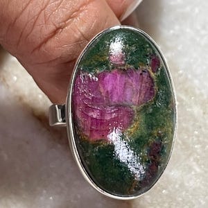 May include: An oval-shaped ring featuring a polished cabochon stone with a vibrant mix of deep magenta-pink ruby inclusions set within a dark green zoisite matrix. The stone is encased in a smooth, polished silver-toned metal bezel setting.
