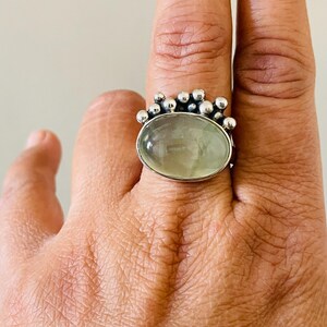 May include: A silver ring featuring a large, oval, pale green gemstone. The ring's band is adorned with small, round silver beads around the gemstone. The ring is displayed on a hand.