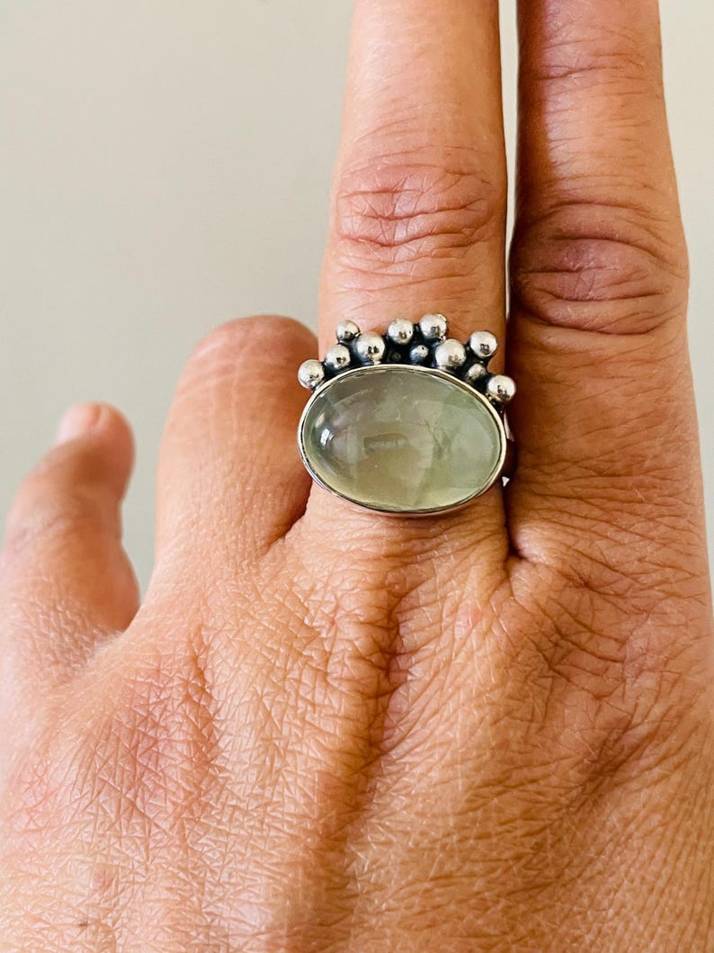May include: A silver ring featuring a large, oval, pale green gemstone. The ring's band is adorned with small, round silver beads around the gemstone. The ring is displayed on a hand.