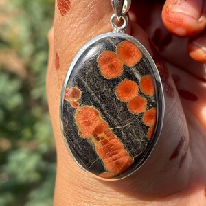 May include: Oval pendant necklace featuring a dark gray stone with vibrant orange spots and patterns. The stone is set in a silver-colored bezel and hangs from a delicate silver chain. A unique piece of jewelry.
