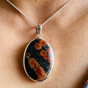 May include: Oval pendant necklace featuring a black stone with orange and red patterns, set in a silver-colored metal frame. The pendant hangs from a delicate silver chain, creating a striking contrast.