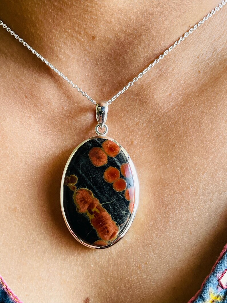 May include: Oval pendant necklace featuring a black stone with orange and red patterns, set in a silver-colored metal frame. The pendant hangs from a delicate silver chain, creating a striking contrast.