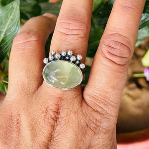 May include: A silver ring featuring an oval, pale green gemstone. The ring has a decorative crown-like design with small, round silver accents surrounding the gemstone. The ring is shown on a hand with a natural background.