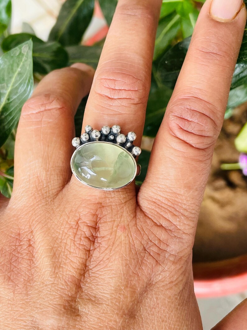 May include: A silver ring featuring an oval, pale green gemstone. The ring has a decorative crown-like design with small, round silver accents surrounding the gemstone. The ring is shown on a hand with a natural background.