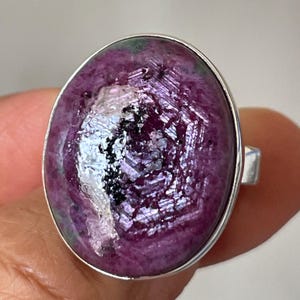 May include: An oval-shaped ring featuring a large, polished cabochon stone with a deep magenta and purple hue, accented by dark green inclusions and a central black crystalline pattern, set within a smooth, polished silver-toned metal band.