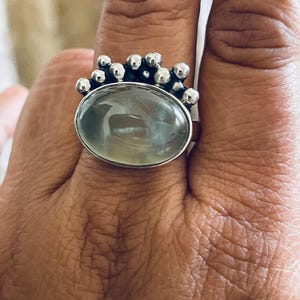 May include: Silver ring featuring a large, oval, pale green gemstone. The ring has a decorative silver band with small, round embellishments clustered around the gemstone. The ring is worn on a finger.