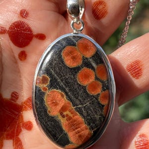 May include: Oval pendant necklace with a silver chain. The pendant features a black stone with orange and tan patterns. The pendant is set in a silver bezel.