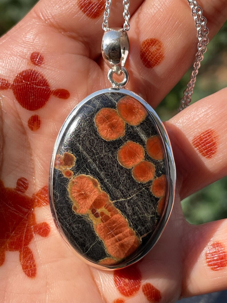 May include: Oval pendant necklace with a silver chain. The pendant features a black stone with orange and tan patterns. The pendant is set in a silver bezel.
